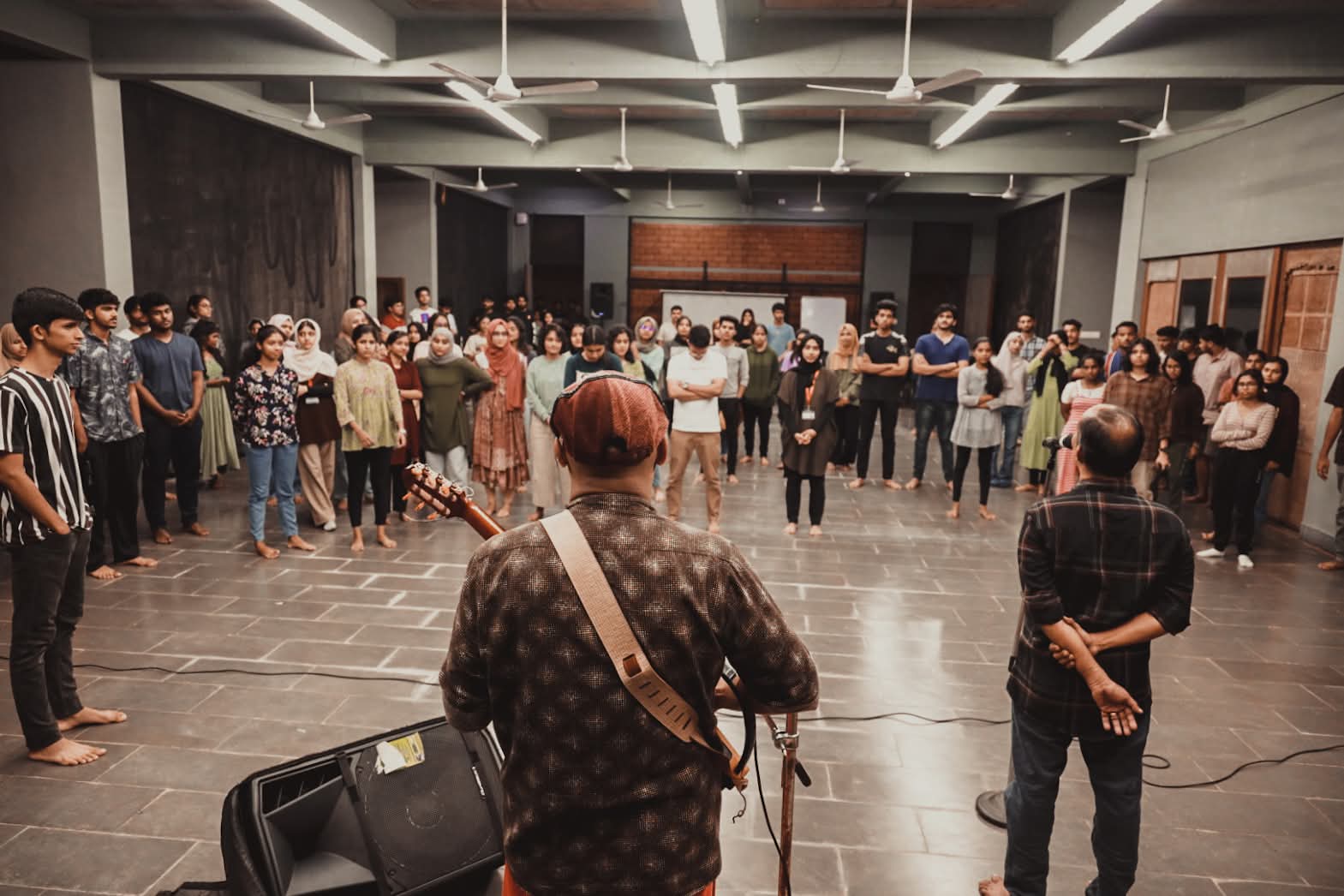 A large circle of students standing barefoot in a college hall around a facilitator with a guitar.