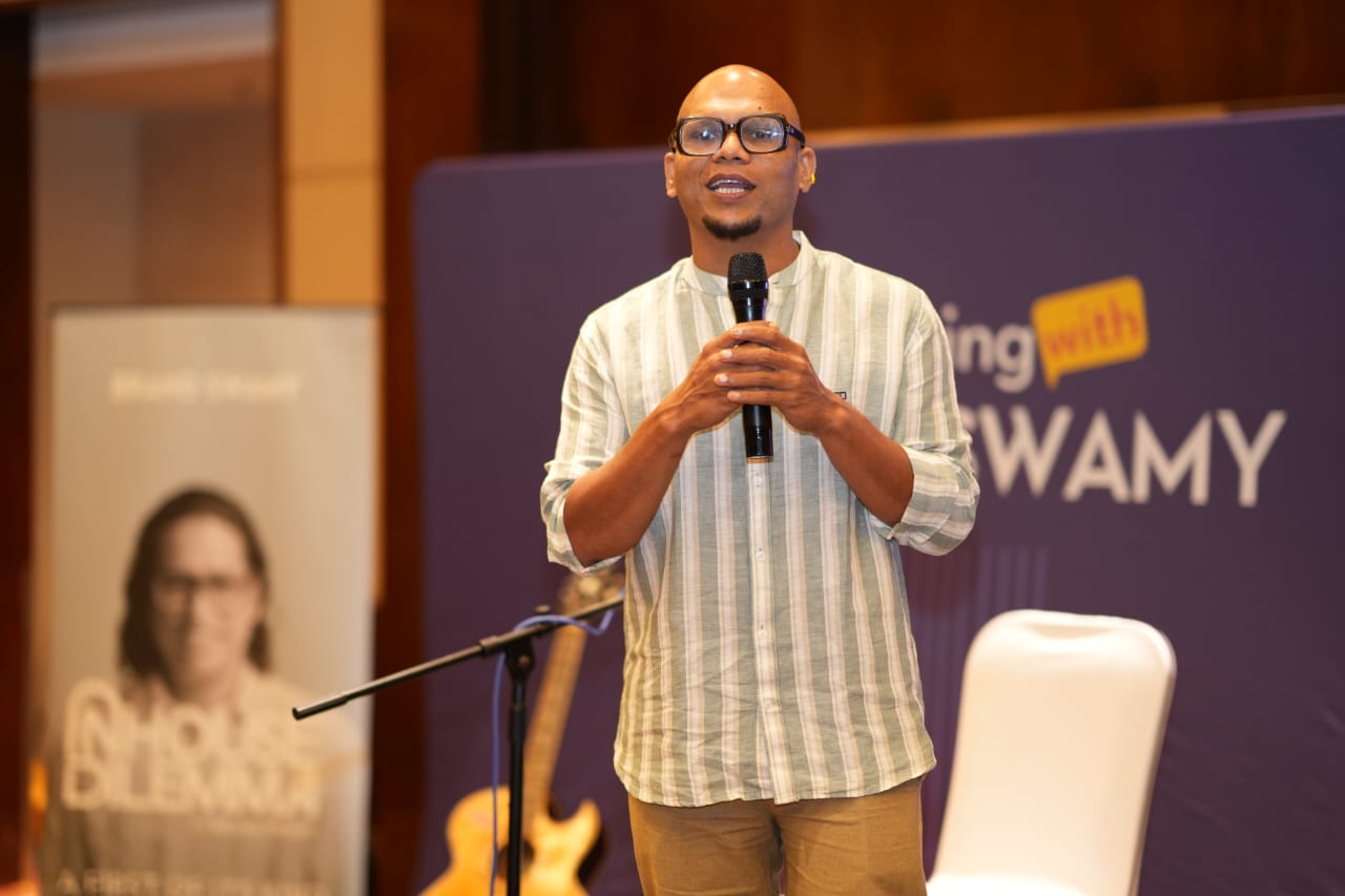 Saji Oorali speaking into a handheld microphone on a stage at a corporate event.